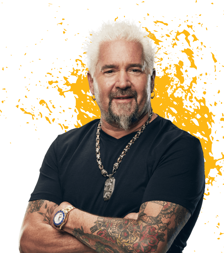 Guy Fieri looking at camera with arms crossed, wearing a black t-shirt, chunky necklace and gold watch